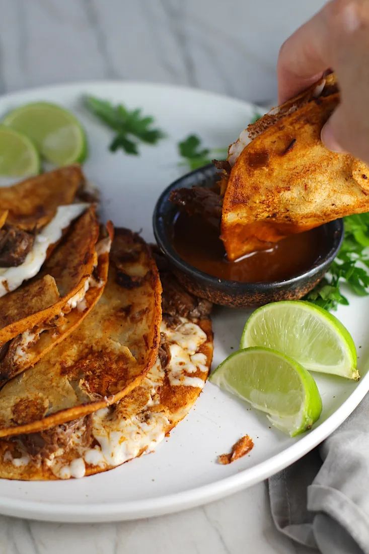 Pressed birria tacos on a white plate, one being dipped into a bowl of consommé, with lime wedges alongside