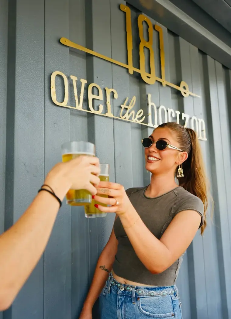Two friends toasting beneath the gold Over the Horizon sign